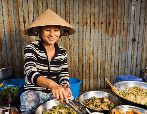 Woman in conical hat serving food from bowls at a market stall.