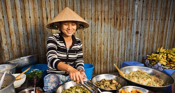 Woman in conical hat serving food from bowls at a market stall.