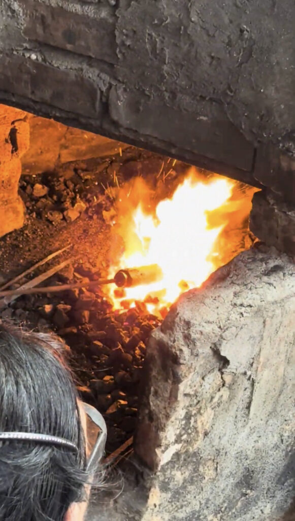 Person working with fire in a forge, with glowing flames and tools visible.