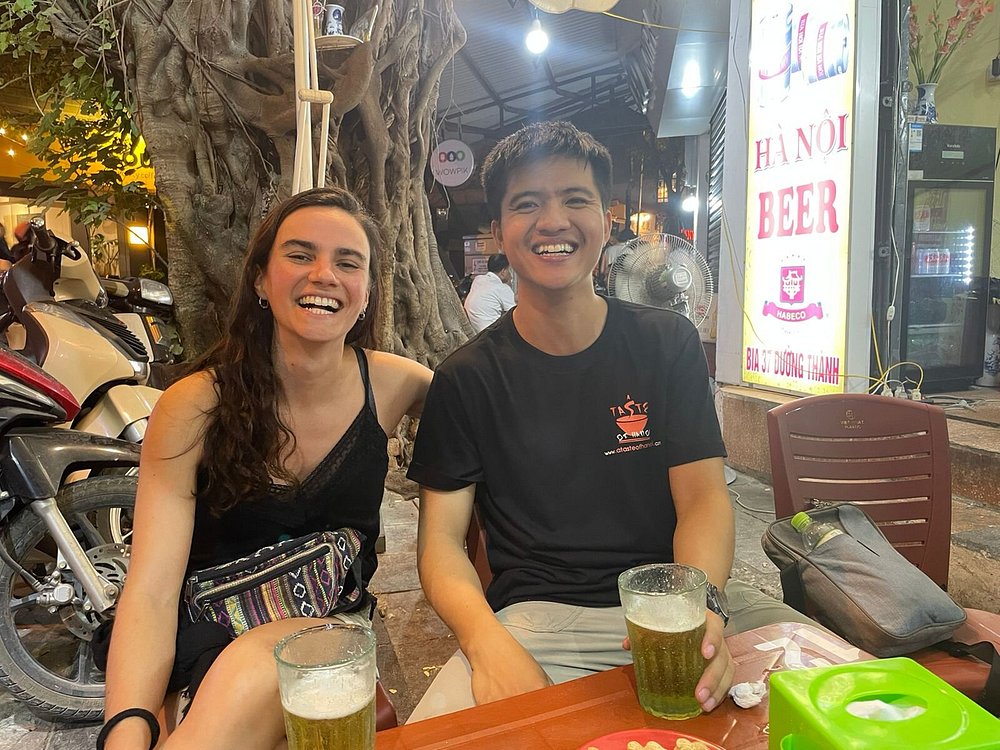 Two people smiling at an outdoor cafe table with drinks and a scooter in the background.