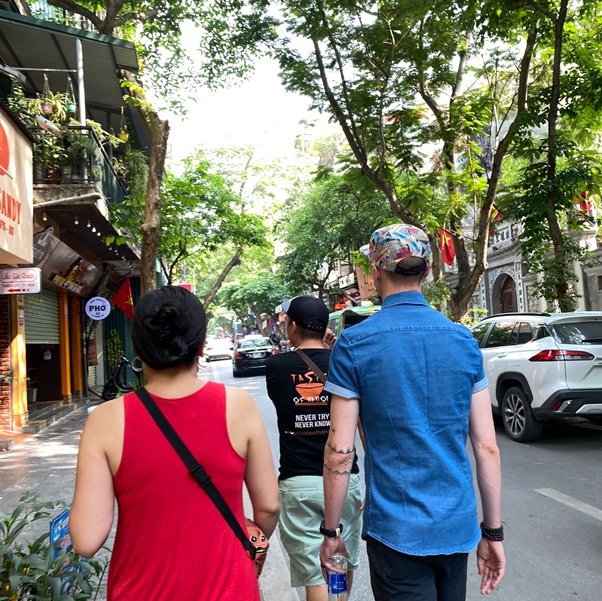 Three people walking on a tree-lined street with cars in Vietnam.