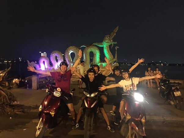 Four people on scooters posing with a lit dragon sculpture at night.