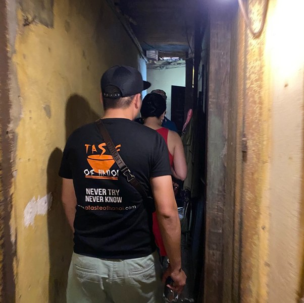 Two people walk through a narrow, dimly lit hallway with peeling walls.