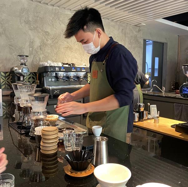 Barista in mask prepares coffee with drip filter at a counter.