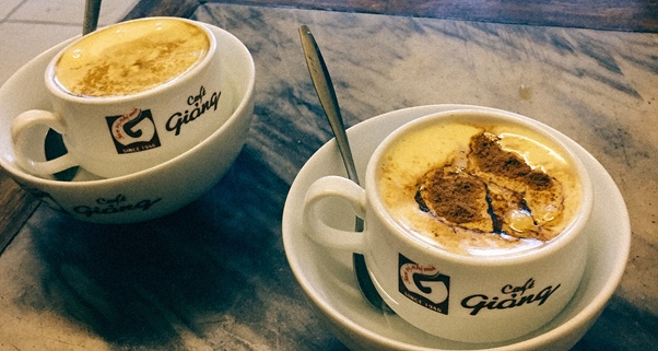 Two cups of coffee with cream froth beside metal spoons on a marble table.