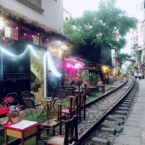 Outdoor café seating along railway in urban street with neon signs and trees.