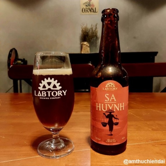 Beer bottle labeled 'SA HUYNH' and a glass with 'LABTORY' logo on a wooden table.