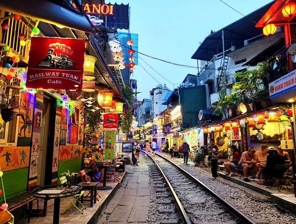 Vibrant street with cafes alongside railway tracks, colorful lights and signs, people sitting and walking.