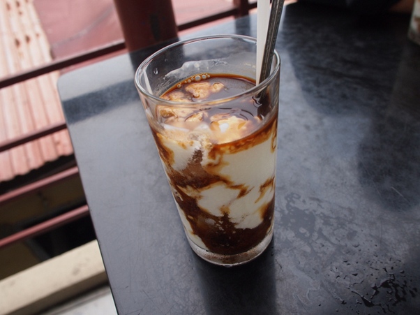 Glass of yogurt with brown syrup swirls and a spoon on a black table.