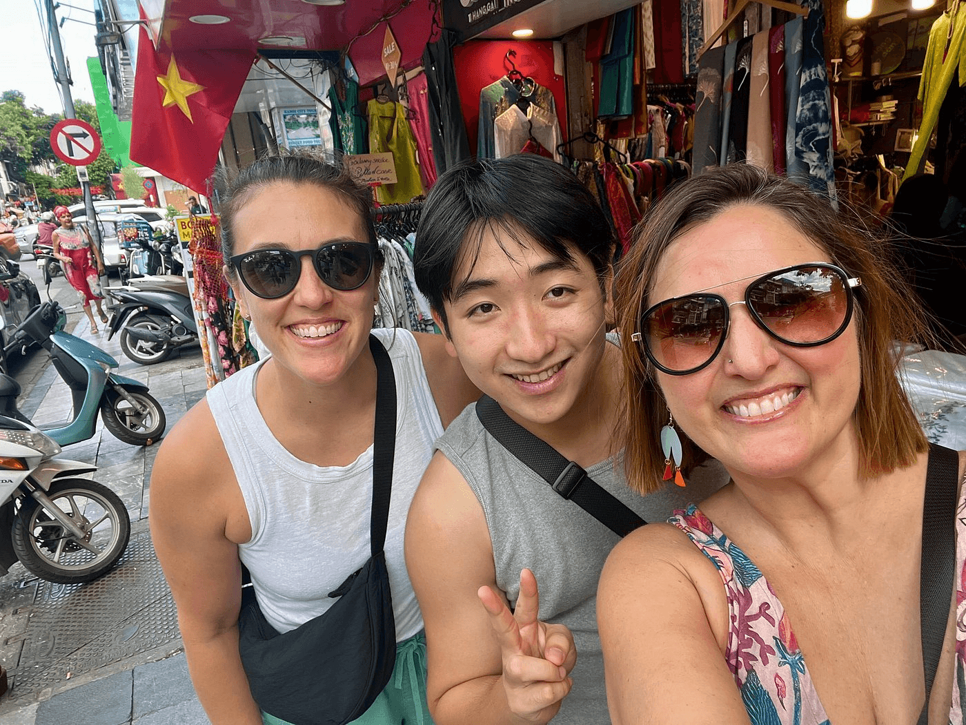 Three people smiling, posing for a selfie on a bustling street with shops and scooters.