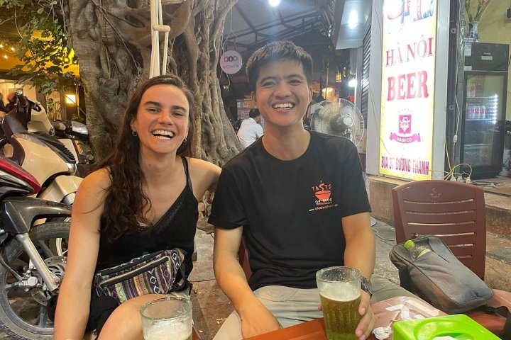 Two people smiling at an outdoor cafe table with drinks and a scooter in the background.
