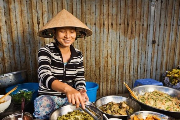 Woman in conical hat serving food from bowls at a market stall.