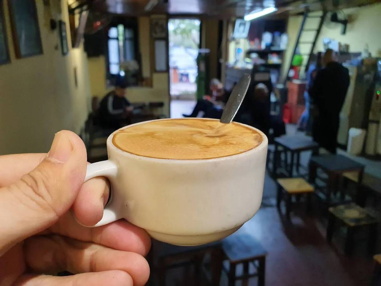 Hand holding a cup of cappuccino with a spoon inside, in a cozy coffee shop.