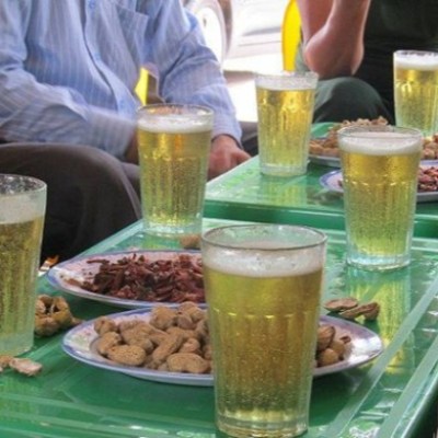 Glasses of beer and plates of snacks on green tables with people sitting nearby.