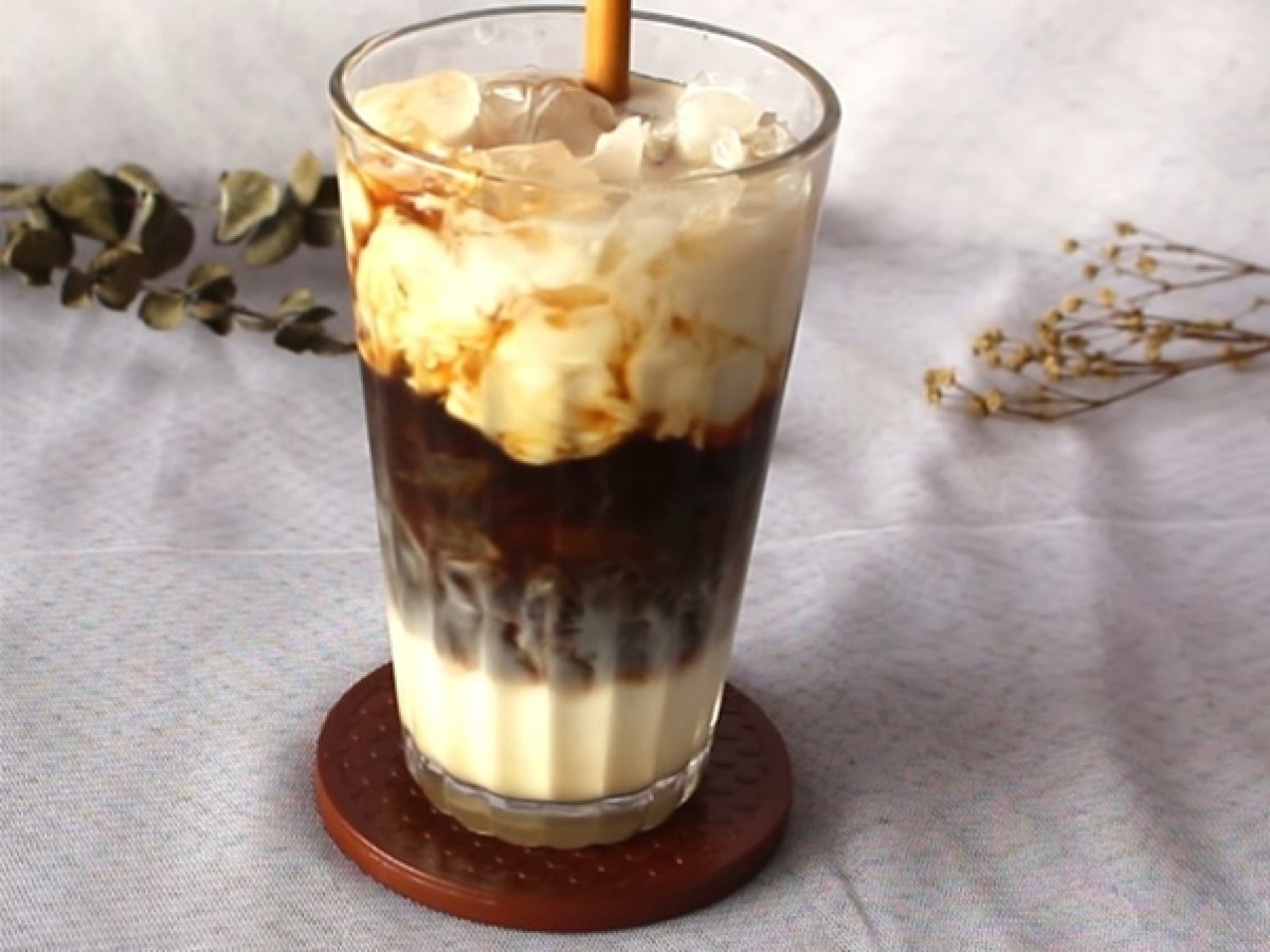 Iced layered coffee with milk in a glass, on a coaster, and a straw on a light background.