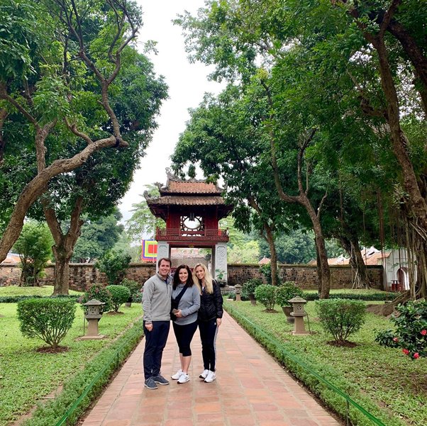Three people stand on a pathway in a garden with a traditional pagoda in the background.