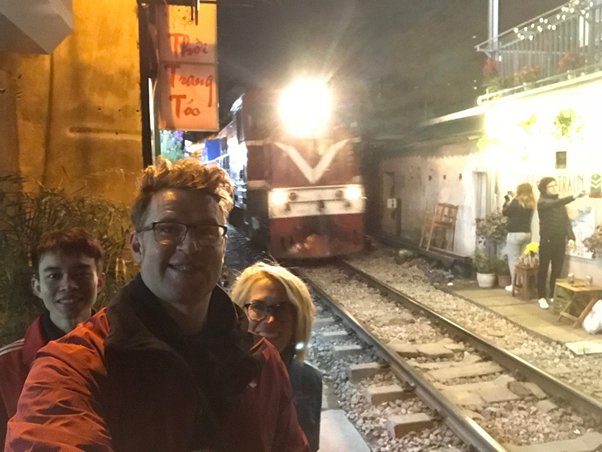 Three people take a selfie near a railway track with an oncoming train at night.
