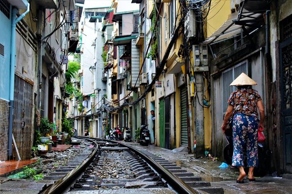 Narrow street with railway track, woman in conical hat walking, lined by closely built buildings.