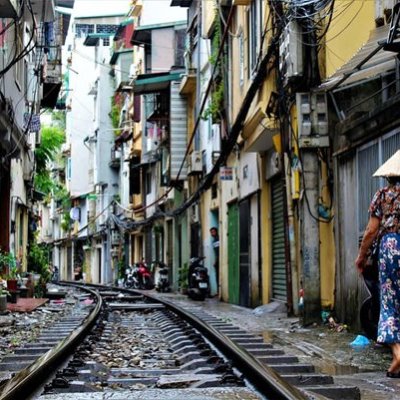Narrow street with railway track, woman in conical hat walking, lined by closely built buildings.