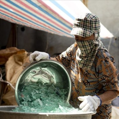 Person wearing protective gear handling broken glass in a metal container under a striped canopy.