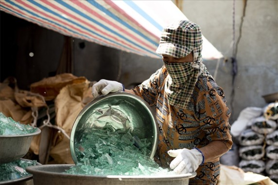 Person wearing protective gear handling broken glass in a metal container under a striped canopy.