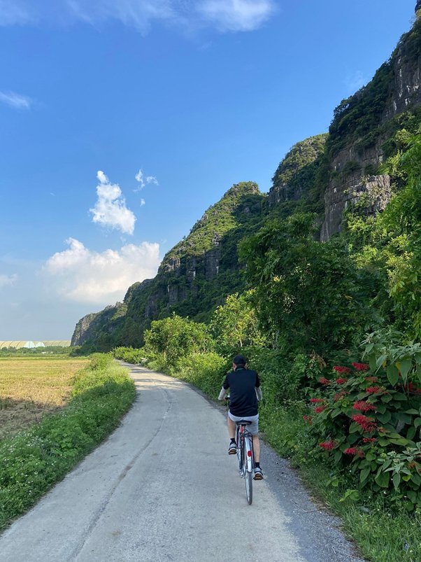 Person cycling on a rural road with green mountains and blue sky.