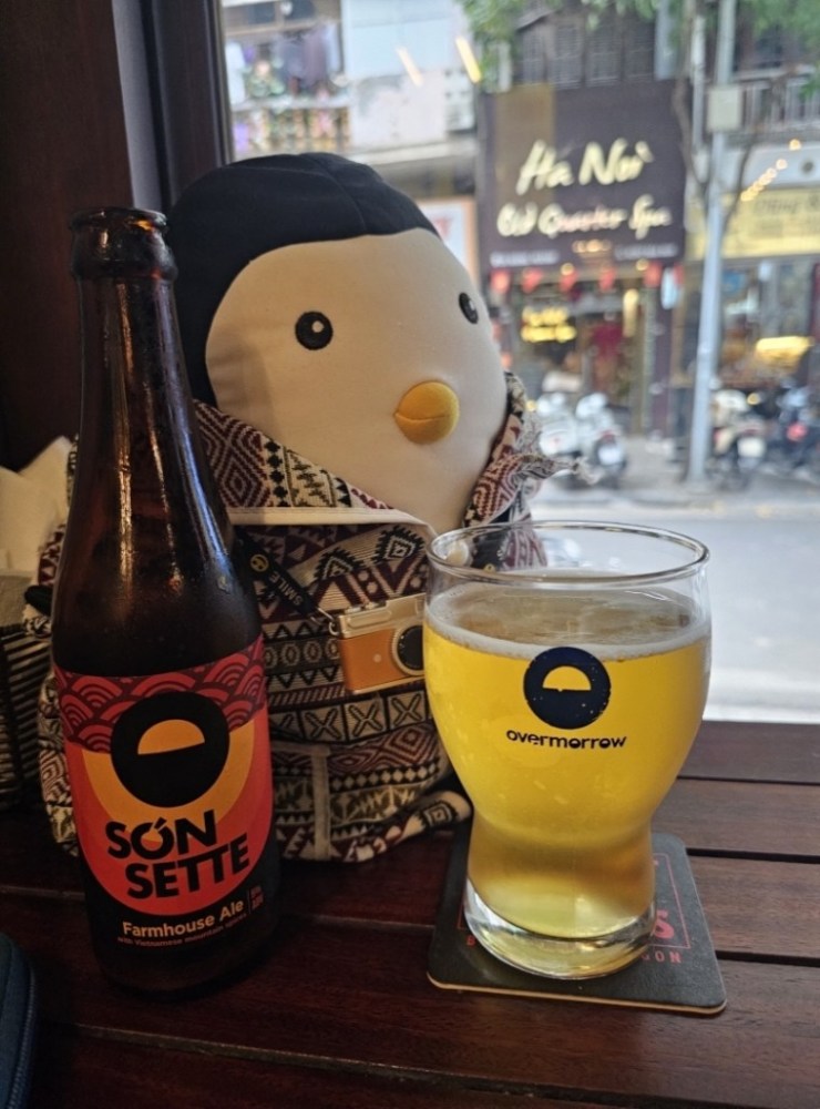 Plush penguin with patterned backpack next to a Són Sette beer bottle and glass on a table.