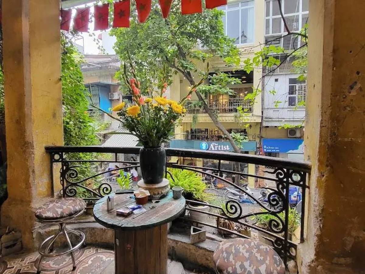 Cozy balcony with round table, two stools, and potted flowers overlooking a street.