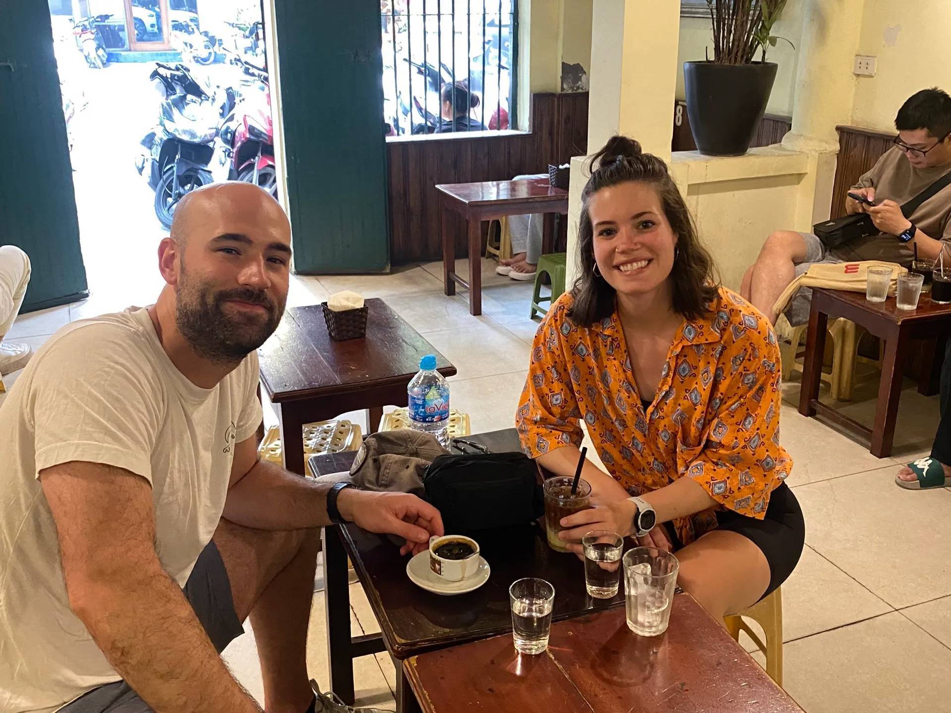 Two people sitting at a café table with drinks, smiling at the camera.