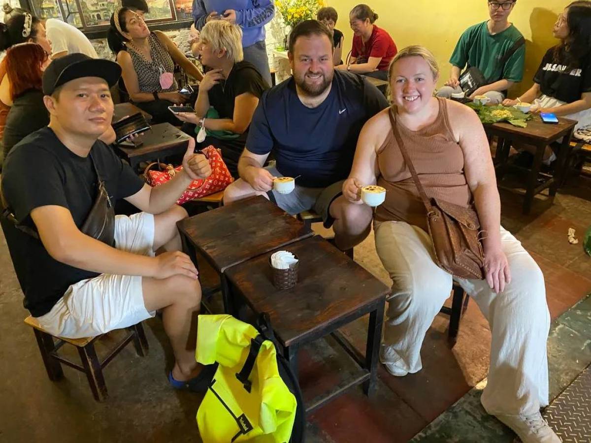 Three people at a cafe table smiling with drinks; others are seated in the background.