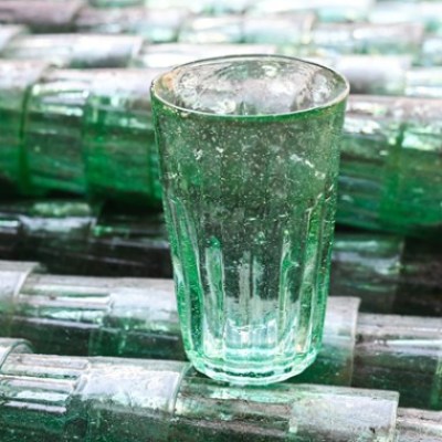A single transparent glass tumbler on stacked green glass bottles.