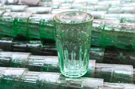 A single transparent glass tumbler on stacked green glass bottles.
