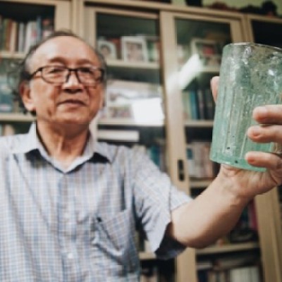 Elderly man in glasses holding a glass, standing in front of book-filled shelves.