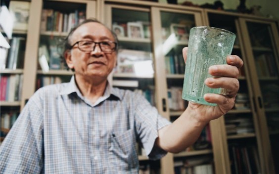 Elderly man in glasses holding a glass, standing in front of book-filled shelves.