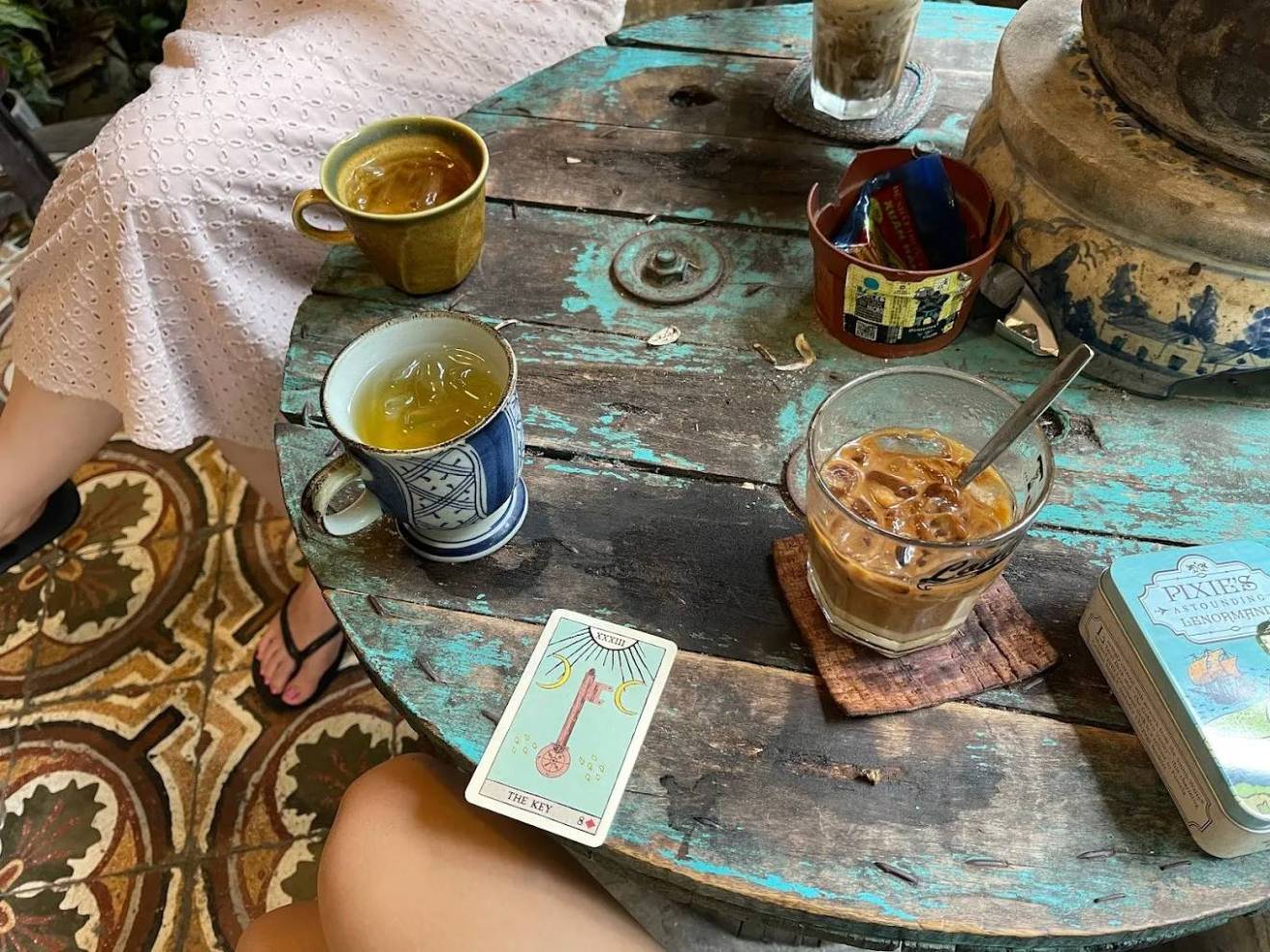 Two iced drinks and a tarot card on a rustic wooden table, with a person's legs visible.