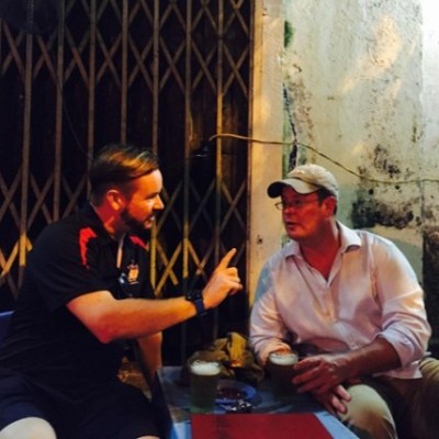 Two men sitting outdoors at a small table with drinks, engaged in conversation.