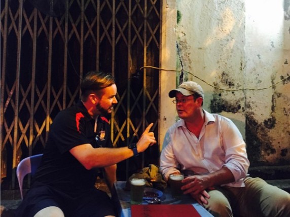 Two men sitting outdoors at a small table with drinks, engaged in conversation.