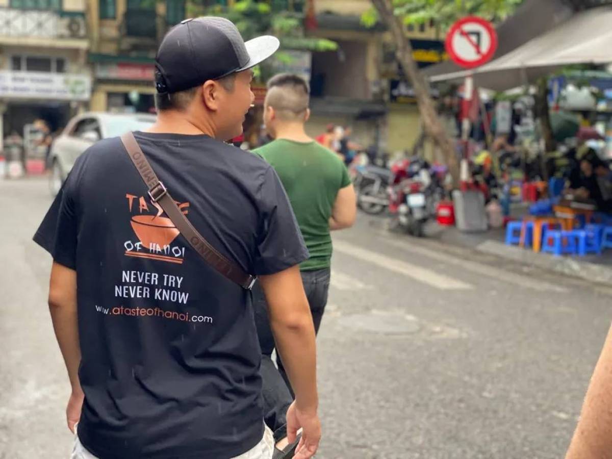 Two men walking on a street; one wears a 'Taste of Hanoi' shirt with a cap.