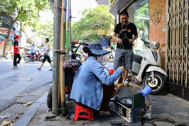 Street vendor seated on a red stool talking to a customer near motorbikes.