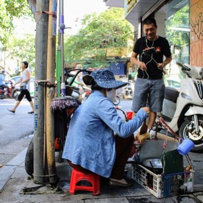 Street vendor seated on a red stool talking to a customer near motorbikes.