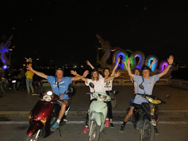 Group of people on scooters at night with colorful dragon sculptures in the background.