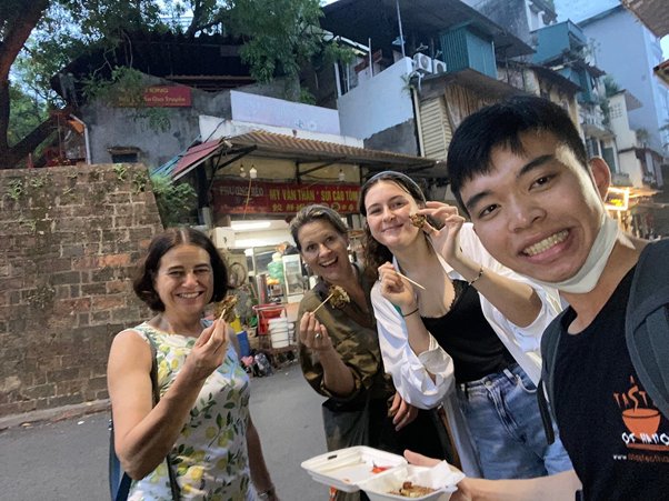 Four people happily pose with food on a lively street.