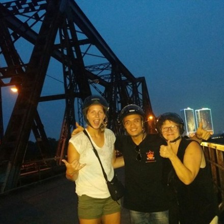 Three people on a bridge at night, two women and one man, smiling with an illuminated skyline in the background.