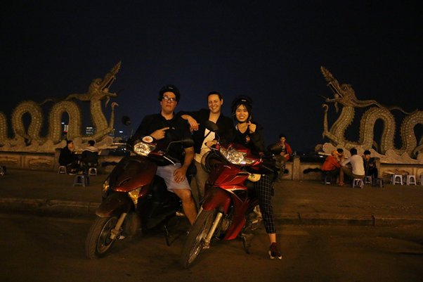 Three people on scooters at night, with dragon statues in the background.