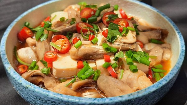 Bowl of sliced mushrooms, tofu, chili peppers, and herbs in sauce.