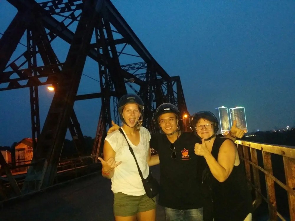 Three people in helmets on a bridge at night, with city lights in the background.