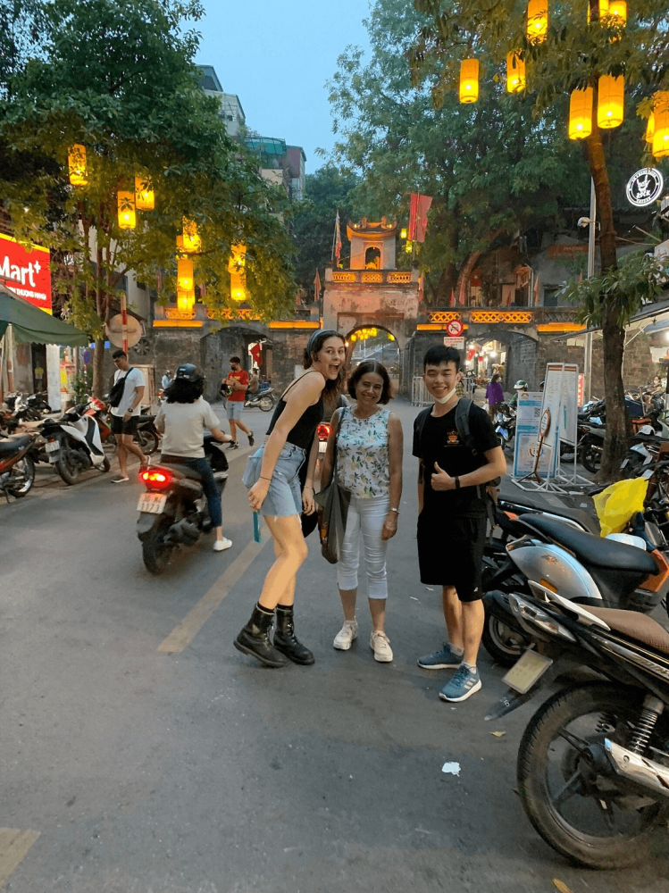 Three people posing on a street with motorbikes and lanterns overhead.