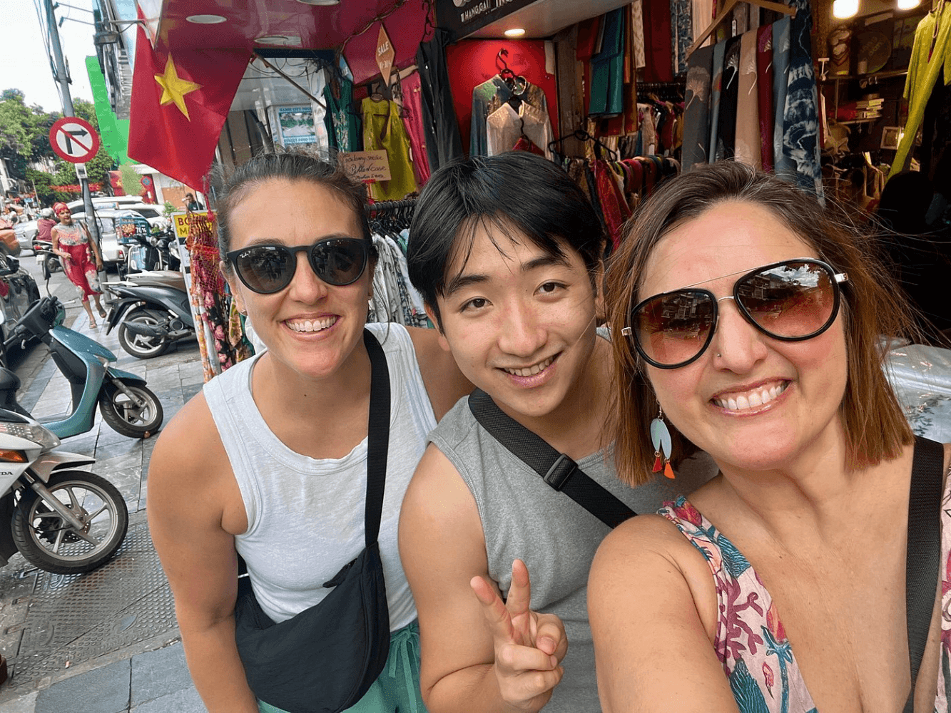 Three people smiling, posing for a selfie on a bustling street with shops and scooters.