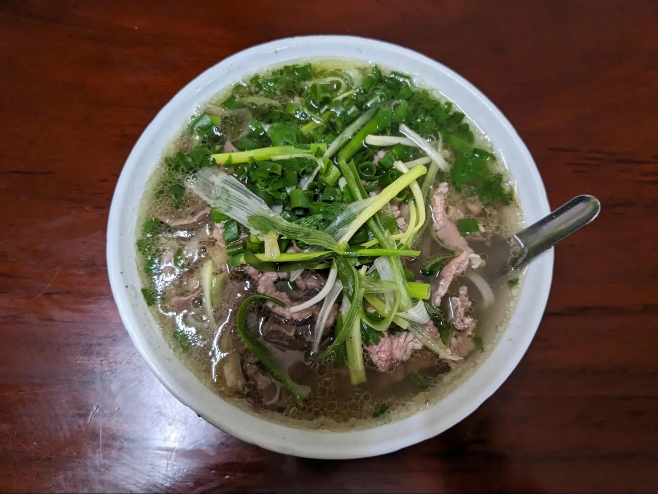 Bowl of pho with greens, beef slices, and a spoon on a wooden table.