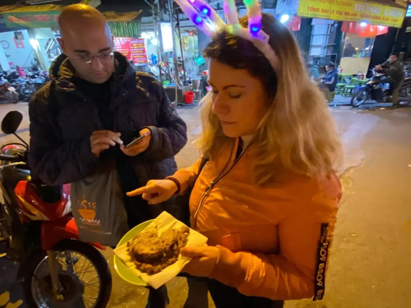 Woman with light-up ears holds food on a street, while man looks at a phone.
